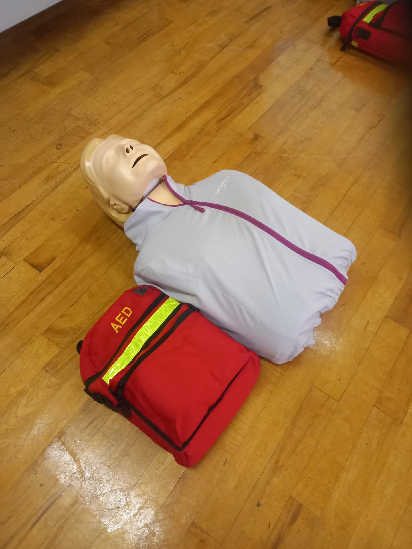 A CPR training manikin and a red AED training bag on a wooden floor